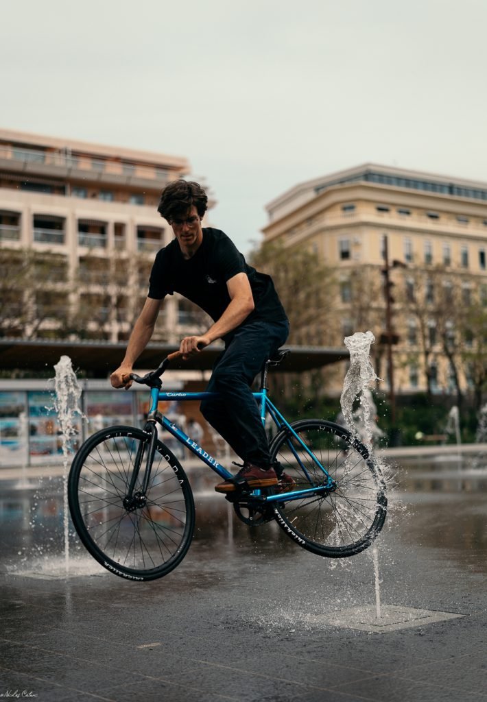 Rider sur un pignon fixe bleu dans l’eau peu profonde d’une place.