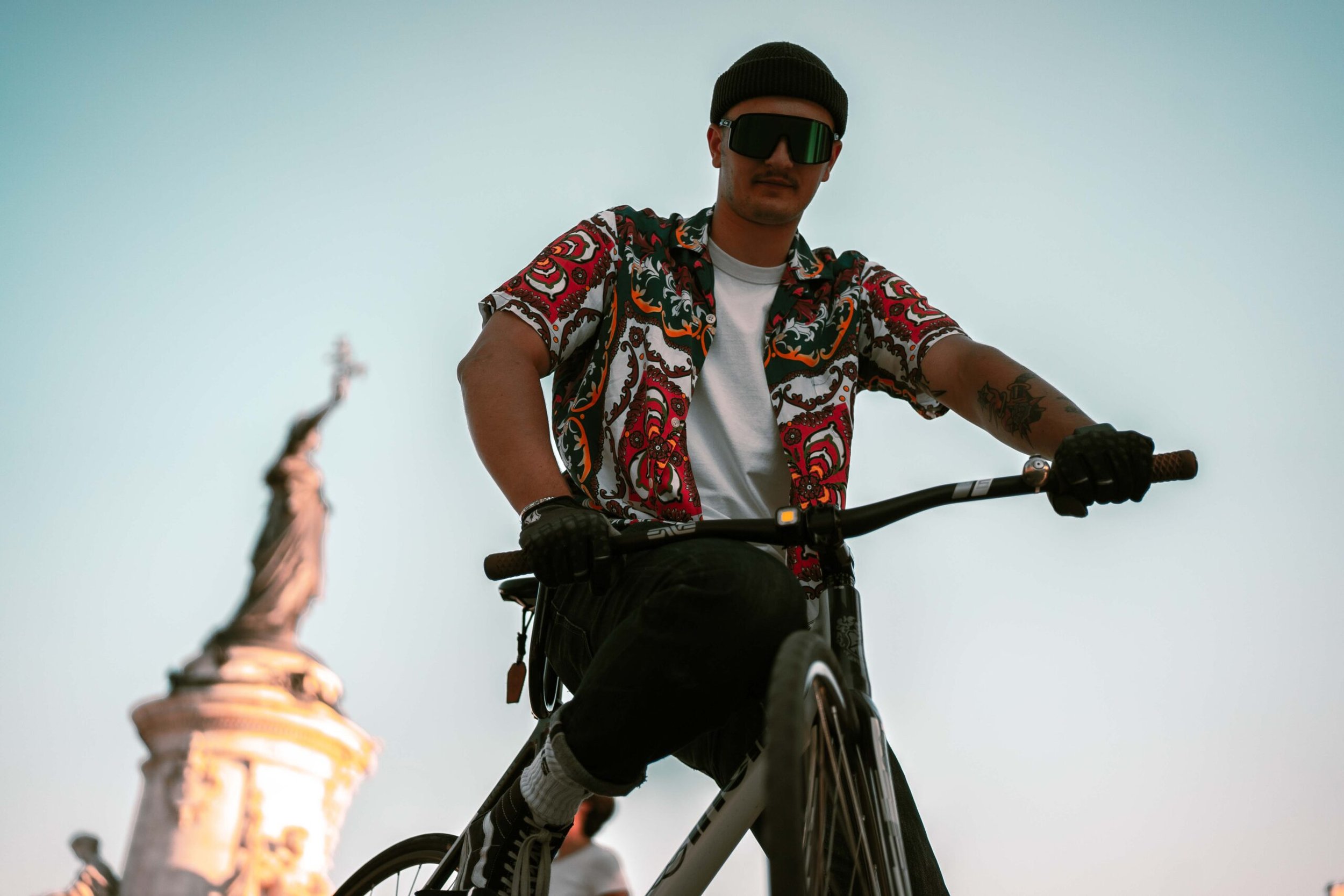 Rider portant des lunettes de soleil et une chemise à motifs, assis sur son vélo sur une place.