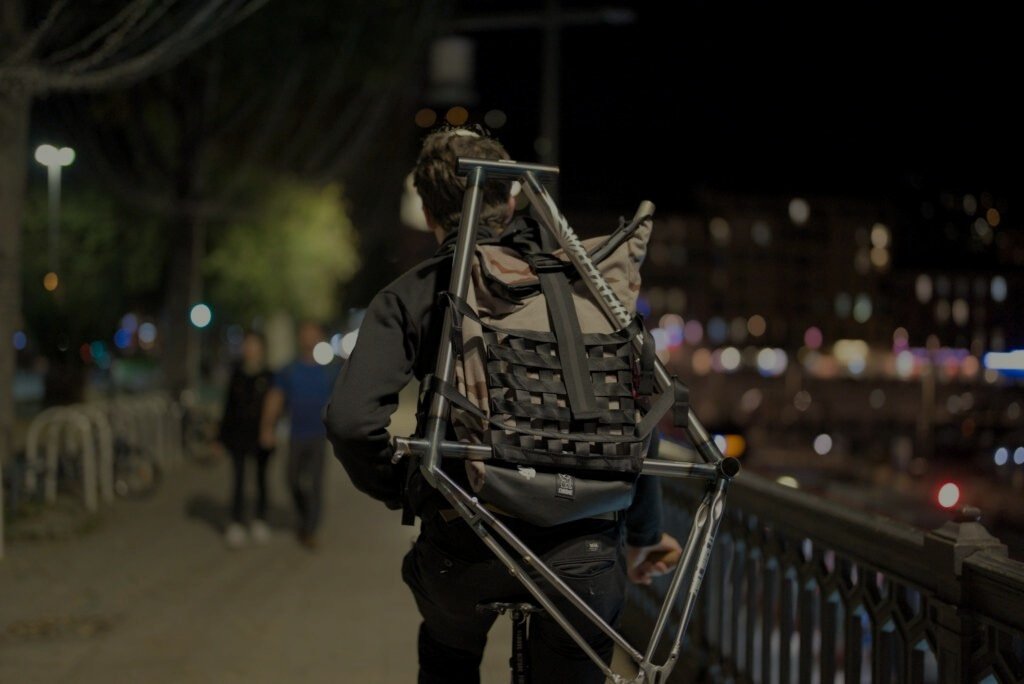 Personne portant un cadre nu et un sac à dos le long d’un garde-corps au bord de l’eau, de nuit.